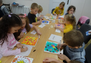 Ośmioro dzieci siedzi przy stole i dokańcza malowanie płótna.